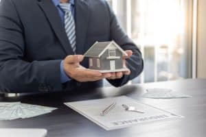A man in a suit holding a model house over a desk with keys and documents