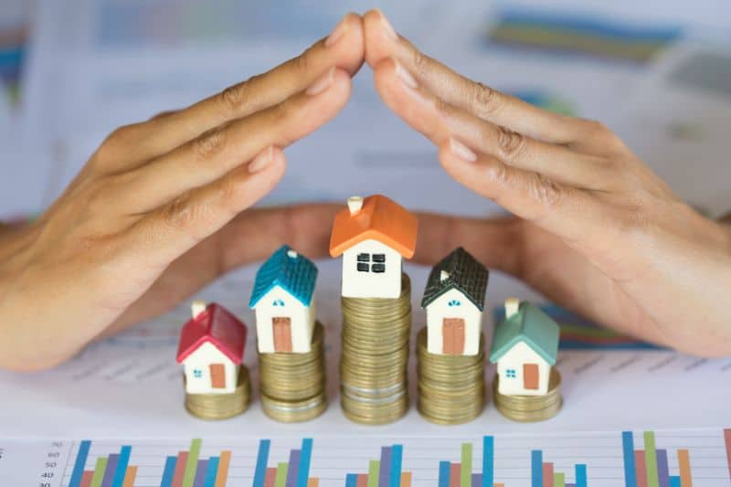 Hands forming a roof over model houses on coin stacks, illustrating investment protection
