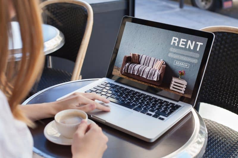 A woman using a laptop displaying the word RENT with a coffee cup